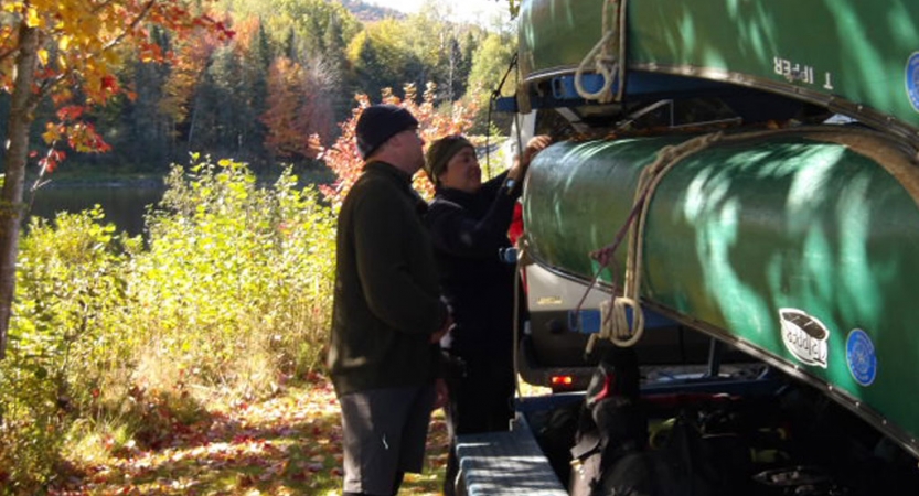 Two people examine canoes stacked on a trailer. Behind them, you can see fall foliage. 